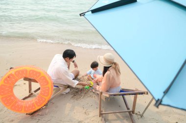 Ebeveynler plaj sandalyelerinde otururken gün içinde kumsalda şemsiye altında küçük çocuklarıyla oynuyorlar.