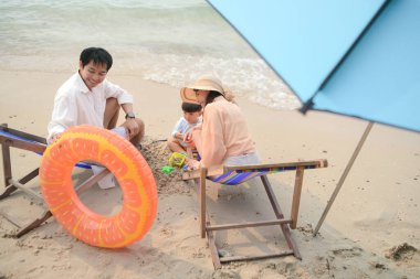 Ebeveynler plaj sandalyelerinde otururken gün içinde kumsalda şemsiye altında küçük çocuklarıyla oynuyorlar.