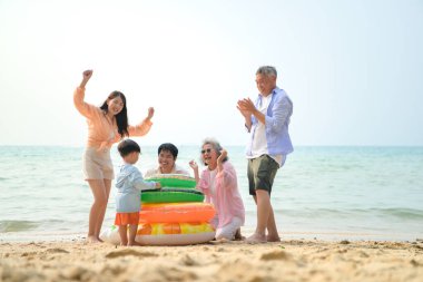 Genişletilmiş aile gündüz vakti deniz kenarında kumlu kumsalda ayakta durup şişme halkalarla oynuyor.