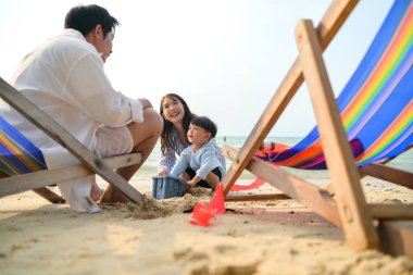 Tatilde, ebeveynler plaj sandalyelerinde oturup, gün boyunca çocuklarının kumsalda kum oyuncaklarıyla oynamasını seyrederler.