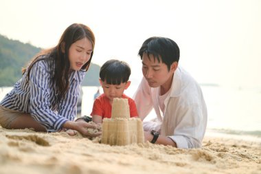 Tatilde, plajda birlikte oynayan genç bir aile, güneşli bir sahil tatili sırasında çocuklarının kumdan kale yapmalarına yardım eden aileler.