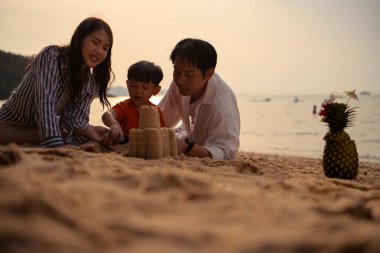 Tatilde, plajda birlikte oynayan genç bir aile, güneşli bir sahil tatili sırasında çocuklarının kumdan kale yapmalarına yardım eden aileler.