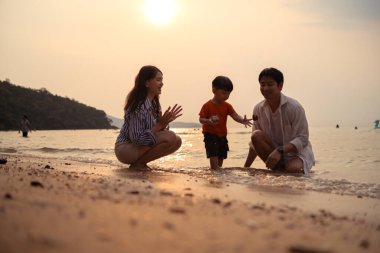 Ebeveynler gün batımında sahilde sığ deniz suyunda yürürken küçük çocuklarıyla el ele tutuşuyorlar.