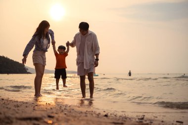 Ebeveynler gün batımında sahilde sığ deniz suyunda yürürken küçük çocuklarıyla el ele tutuşuyorlar.