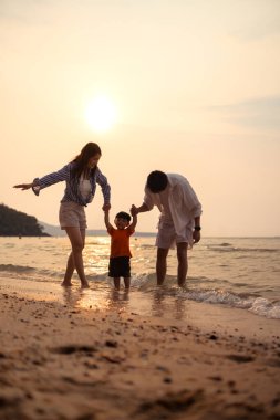Ebeveynler gün batımında sahilde sığ deniz suyunda yürürken küçük çocuklarıyla el ele tutuşuyorlar.