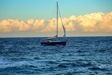 Gün batımında açık denizde süzülen parlak yelkenli altın bulutlar ve canlı mavi sularla çerçevelenmiş. Yolculuğun, maceranın, özgürlüğün ve yelkenciliğin güzelliğinin mükemmel bir görüntüsü..