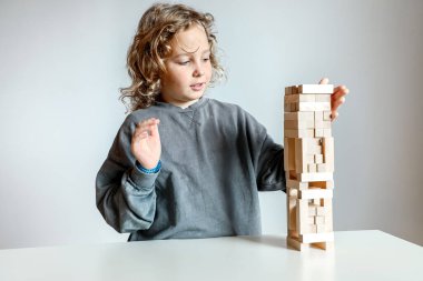 Düşünceli kıvırcık saçlı çocuk beyaz bir masanın üzerine tahta blok bir yapı inşa ediyor. Odaklanmayı, hayal gücünü ve oyun oynayarak neşeli öğrenmeyi sembolize ediyor..