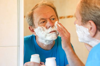 Morning light reflects off the mirror as a man spreads shaving foam on his cheeks, creating a calm moment of self-care. This scene evokes a familiar, comforting start to the day.