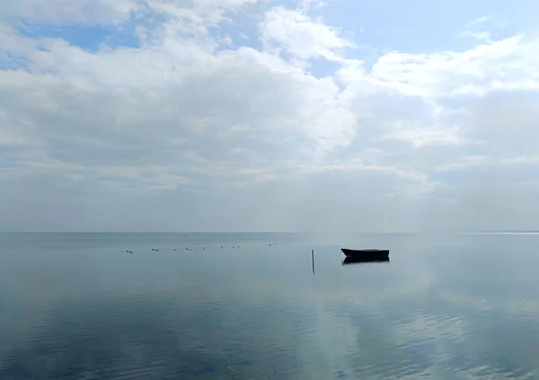 Lonely boat floating on calm sea under soft daylight and cloudy sky, minimalistic seascape with reflections, peaceful horizon view, tranquil nature background, no people, serene coastal concept.