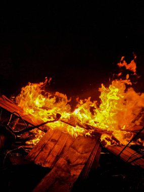 Parlak alevlerin bir odun yığını üzerinde dans ettiği parlak bir ateşin görüntüsü. Koyu bir zemin karşısında turuncu ve sarı alevlerin karşıtlığı yoğun bir atmosfer yaratır..