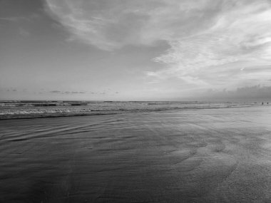 Şafak vakti kumsalın sakin bir görüntüsü, siyah ve beyaz, yumuşak dalgalar tarafından ele geçirilmiş ıslak kumu ve kıyıda yürüyen uzak insanları..