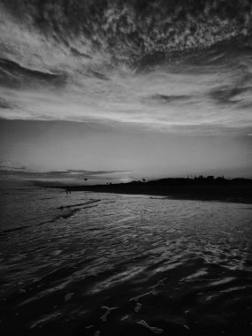 Gün batımında kumsalda, siyah ve beyazla kaplanmış, bulutlarla kaplı dramatik bir gökyüzünü ve sudaki yansımalarını gözler önüne seriyor. Uzak insanların siluetleri huzur ve tefekkür katar..