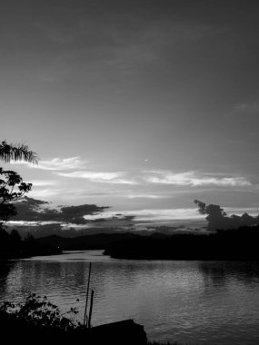 Bir nehrin üzerinde sakin bir gün batımının güzel siyah-beyaz sahnesi, arka planda ağaçların ve dağların siluetleri, sakin ve düşünceli bir atmosfer yaratıyor..