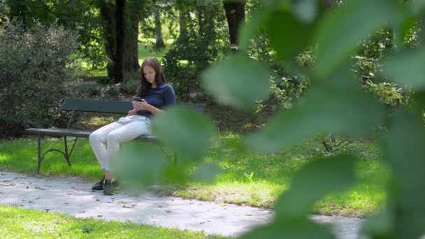 Menina com um telefone celular em um banco de parque — Vídeo de Stock
