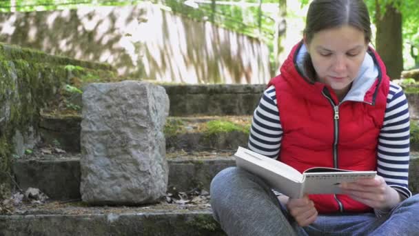 Het meisje in een rode jas en een gestreepte trui, het lezen van een groot boek op een prachtige locatie op de oude trappen. — Stockvideo