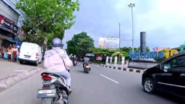 5 Nisan 2025 'te, Bandung, Batı Java, Endonezya' daki iş yerlerine giderken, güneşli bir kavşakta motosiklet ve araba sürenlerin 4K kaydı.