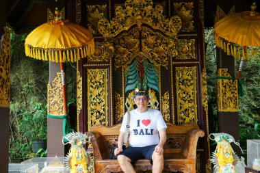 Geleneksel ahşap bir bankta oturan erkek bir turistin güzel bir Bali tapınağı köşkünde altın oymalı bir tişört giyerken fotoğrafı. Bali, 22 Temmuz 2025.