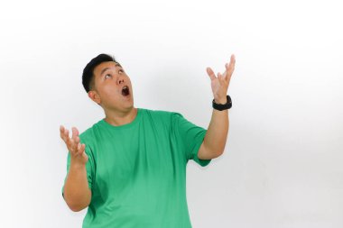 İki eli havada, yeşil tişörtlü, hayret içinde Asyalı bir adam kameraya büyük gözlerle bakıyor ve ağzını açıyor..
