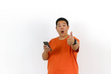 Turuncu tişörtlü heyecanlı Asyalı adam akıllı telefon kullanıyor ve baş parmak işareti yapıyor, tatmin ve onay veriyor..