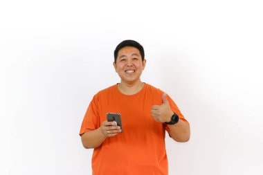 Turuncu tişörtlü heyecanlı Asyalı adam akıllı telefon kullanıyor ve baş parmak işareti yapıyor, tatmin ve onay veriyor..