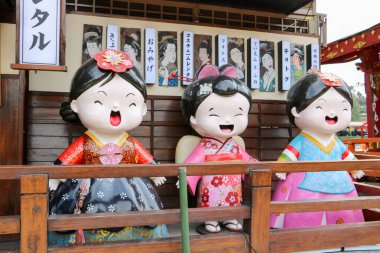 Geleneksel renkli kimonolara bürünmüş üç büyük Japon bebek heykeli ahşap bir çitin arkasında duruyor..