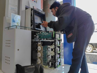 Kahverengi kapüşonlu teknisyen, 1 Ağustos 2025 'te Bali, Endonezya' da yapılan rutin bakım kontrolü sırasında BRI ATM makinesini tamir ediyor.