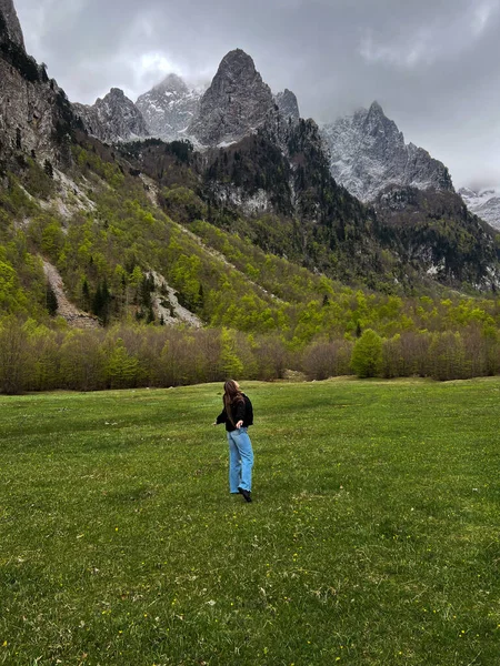 Karadağ 'ın Prokletije Dağları' ndaki dramatik kayalık tepeleri ve bulutlu gökyüzü ile yeşil bir dağ çayırında duran genç kadın.