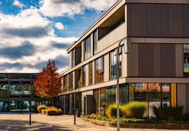 Gilching, Bavyera 'da Modern Mimarlık: Cam Yüzlü Çağdaş Ofis Binası, Sonbahar Ağaçları ve Kentsel İş Bölgesi' nde Dramatik Gökyüzü. Yüksek kalite fotoğraf
