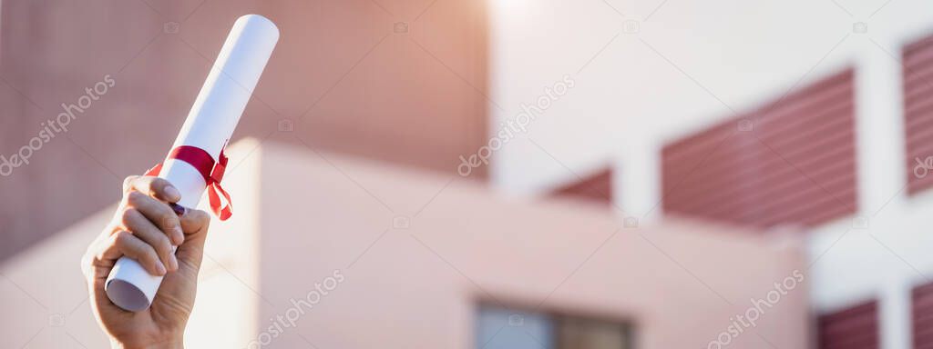 Close-up shot of a university graduate holding degree certification to shows and cerebrate his success in the college commencement day with sunlight in background.