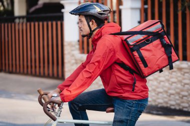 Kırmızı üniformalı kurye bisikletli bir teslimat kutusuyla ve müşteriye yemek teslimatı için adresi kontrol etmek üzere cep telefonuna bakıyor. Bisikletli bir kurye şehre yiyecek getiriyor..