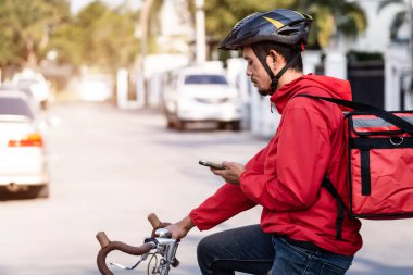 Kırmızı üniformalı kurye bisikletli bir teslimat kutusuyla ve müşteriye yemek teslimatı için adresi kontrol etmek üzere cep telefonuna bakıyor. Bisikletli bir kurye şehre yiyecek getiriyor..