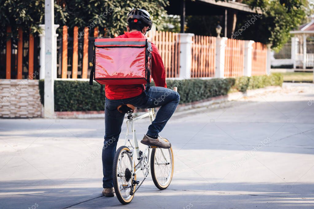 Courier in red uniform with a delivery box on back riding a bicycle and looking on the cellphone to check the address to deliver food to the customer. Courier on a bicycle delivering food in the city.