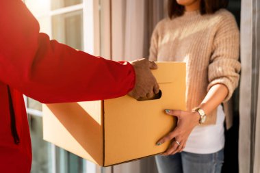 Kırmızı üniformalı Asyalı bir teslimatçı evin önünde kadın bir müşteriye paket teslim ediyor. Bir postacı ve ekspres teslimat servisi covid19 salgını sırasında paket teslim etti..