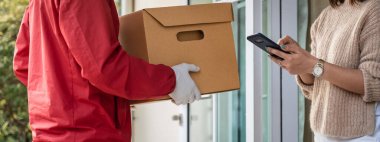 Kırmızı üniformalı Asyalı bir teslimatçı evin önünde kadın bir müşteriye paket teslim ediyor. Bir postacı ve ekspres teslimat servisi covid19 salgını sırasında paket teslim etti..