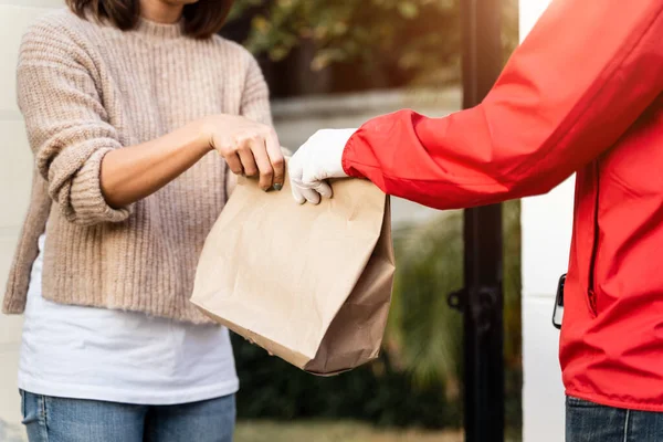 Kırmızı üniformalı Asyalı bir teslimatçı evin önünde kadın bir müşteriye paket teslim ediyor. Bir postacı ve ekspres teslimat servisi covid19 salgını sırasında paket teslim etti..