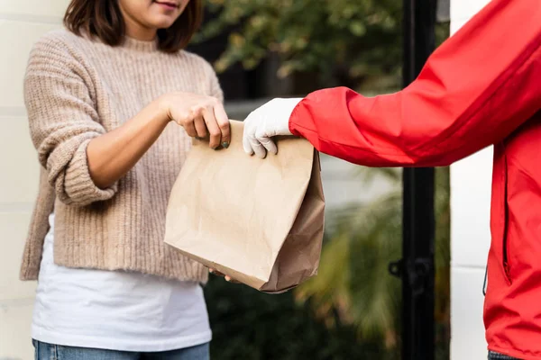 Kırmızı üniformalı Asyalı bir teslimatçı evin önünde kadın bir müşteriye paket teslim ediyor. Bir postacı ve ekspres teslimat servisi covid19 salgını sırasında paket teslim etti..