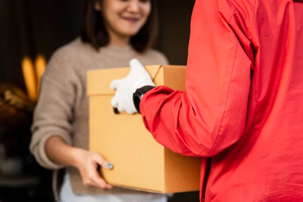 An Asian deliver man in red uniform handing parcel to a female costumer in front of the house. A postman and express delivery service deliver parcel during covid19 pandemic.