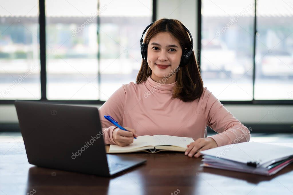 Young asian girl student wears wireless headphones write on the notebook to study language online watch and listen to the lecturer, webinar via video call e-learning at home, distance education