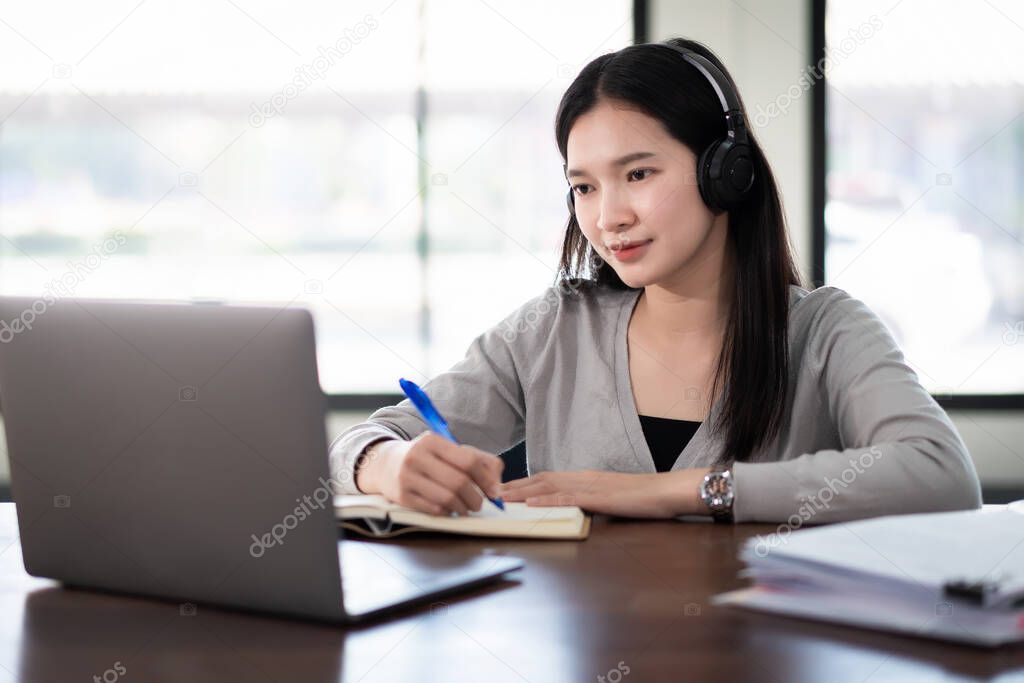 Young asian girl student wears wireless headphones write on the notebook to study language online watch and listen to the lecturer, webinar via video call e-learning at home, distance education