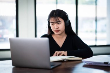 Gülümseyen Asyalı kız öğrenci kablosuz kulaklık takıyor. İnternette dil eğitimi almak ve hocayı dinlemek için deftere yazı yazıyor. Evdeki e-öğrenme videosuyla Webinar, uzaktan eğitim.