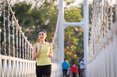 Spor kıyafetleri içinde koşan genç bir Asyalı kadın sporcu sabah şehir parkında antrenman yapıyor. Tropik parkta koşan sağlıklı, genç, sportif, Asyalı bir kadın. Spor ve eğlence.