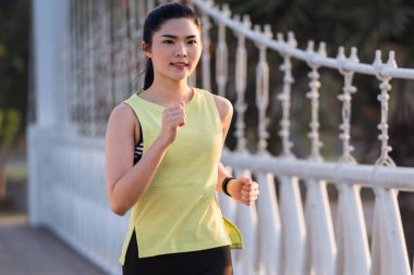 Spor kıyafetleri içinde koşan genç bir Asyalı kadın sporcu sabah şehir parkında antrenman yapıyor. Tropik parkta koşan sağlıklı, genç, sportif, Asyalı bir kadın. Spor ve eğlence.