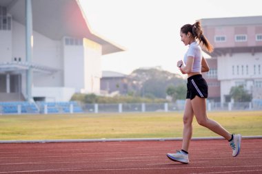 Genç bir Asyalı bayan atlet, formda ve sağlıklı yaşam tarzını korumak için güneşli bir sabahta şehir stadyumunda koşu pistinde koşuyor. Genç fitness kadını stadyum pistinde koşuyor. Spor ve eğlence.