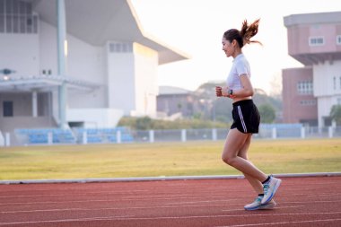 Genç bir Asyalı bayan atlet, formda ve sağlıklı yaşam tarzını korumak için güneşli bir sabahta şehir stadyumunda koşu pistinde koşuyor. Genç fitness kadını stadyum pistinde koşuyor. Spor ve eğlence.