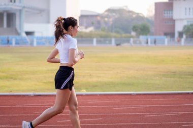 Genç bir Asyalı bayan atlet, formda ve sağlıklı yaşam tarzını korumak için güneşli bir sabahta şehir stadyumunda koşu pistinde koşuyor. Genç fitness kadını stadyum pistinde koşuyor. Spor ve eğlence.