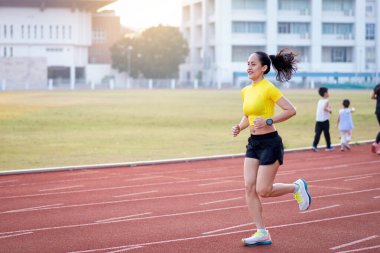 Genç bir Asyalı bayan atlet, formda ve sağlıklı yaşam tarzını korumak için güneşli bir sabahta şehir stadyumunda koşu pistinde koşuyor. Genç fitness kadını stadyum pistinde koşuyor. Spor ve eğlence.