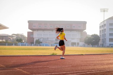 Genç bir Asyalı bayan atlet, formda ve sağlıklı yaşam tarzını korumak için güneşli bir sabahta şehir stadyumunda koşu pistinde koşuyor. Genç fitness kadını stadyum pistinde koşuyor. Spor ve eğlence.