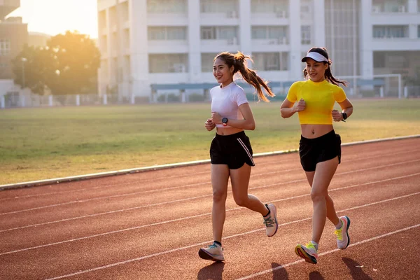 Two young Asian women in sports outfits jogging on running track in city stadium in the sunny morning to keep fitness and healthy lifestyle. Young fitness women run on the stadium track. Sports and recreation