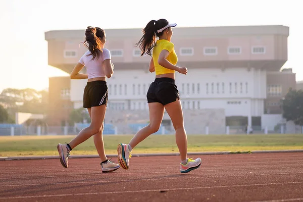 Two young Asian women in sports outfits jogging on running track in city stadium in the sunny morning to keep fitness and healthy lifestyle. Young fitness women run on the stadium track. Sports and recreation