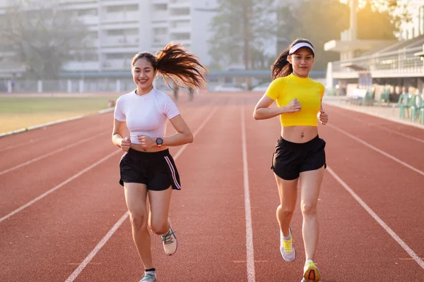 Two young Asian women in sports outfits jogging on running track in city stadium in the sunny morning to keep fitness and healthy lifestyle. Young fitness women run on the stadium track. Sports and recreation